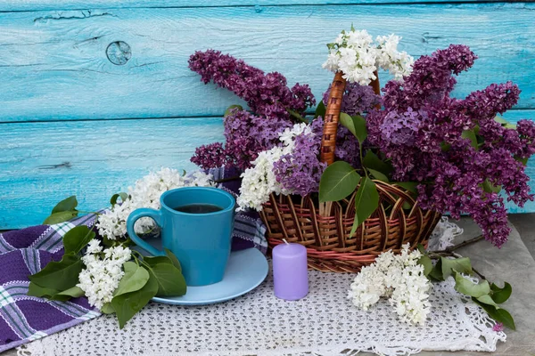 Beyaz bir peçetenin üzerinde bir fincan çay var. Yakınlarda bir sepet içinde beyaz ve mor leylaklar ve bir mum var..