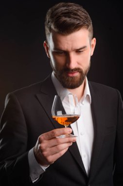 Adam yarı dolu bardaklı. Oturum konyak tatma iyi bilinen bir marka tarafından üretilen. Dikey atış zarif siyah beyaz genç adamın önünde koyu arka plan uygun.