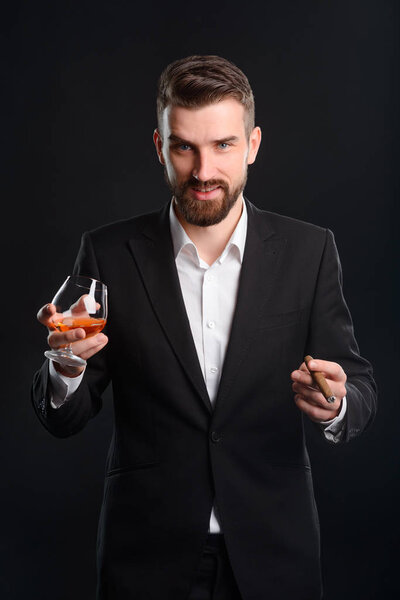 Rich man with drink and cigar. Time of refreshment for a successful businessman. Vertical portrait of man in black suit and white shirt over black background.