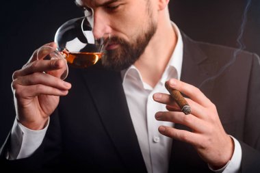 Yudumlarken brendi ve puro sahip. Siyah takım elbise ve unfastened tepe düğme ile beyaz gömlek giymiş genç işadamı sağlam görünümlü görünümünü. Çok az duman Sigara tarih seçici odak.