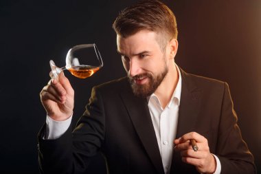 Bir konyak cam tutan adam. Kalitesini değerlendirmek için şık adam siyah takım elbise ve beyaz gömlek güçlü içecek bir bardak arıyorum. Turuncu backlights ve siyah arka plan.