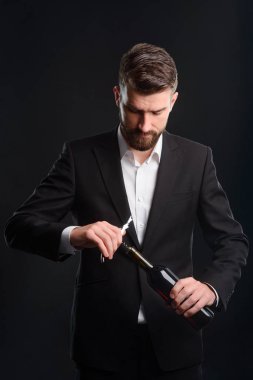Bir şişe kırmızı şarap açma. Şık genç adam bir tirbuşon kullanarak unfastened üst düğme ile siyah takım elbise ve beyaz gömlek giymiş. Sommelier siyah arka plan üzerine dikey portresi.