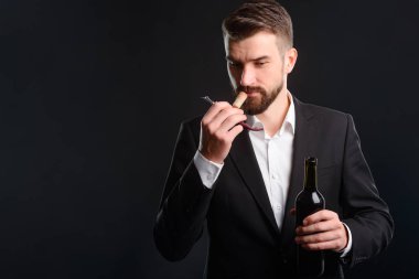 Oturum kırmızı şarap tatma. Biçimsel tarzı kıyafetler giymiş genç adam sadece bir şişe açıldı. Yakışıklı sommelier metninizi eklemek için boş alan ile siyah arka plan üzerine mantar kokulu üzerinde duruldu.
