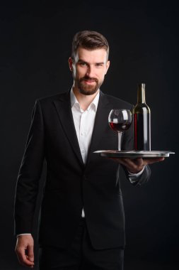 Vertical portrait of wine connoisseur on black background. Bearded man in expensive formal suit looking at the camera while holding a tray with a glass and bottle of red wine. clipart