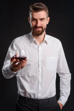 Bir bardağı tutan adam. Beyaz gömlek giymiş ve elinde bir yarısı dolu bardak kırmızı şarap yakışıklı sommelier hafif bir gülümseme. Siyah arka plan ile dikey portre.