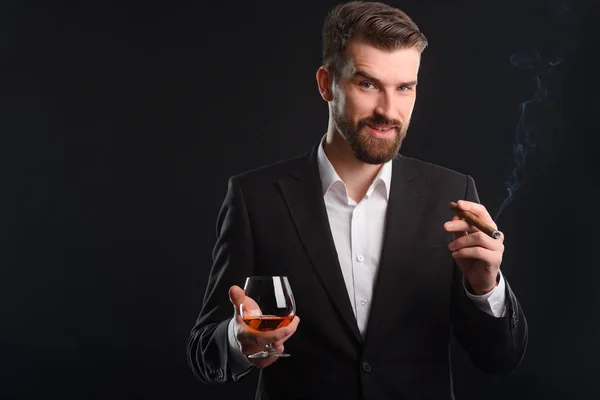 Portrait of solid businessman with black background. Slight smile and uplifted mood of young successful boss. A glass of fine quality brandy and a little fuming cigar in his hands.