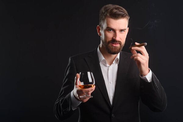 Boss holding cigar and glass. Dark background behind young man with beard looking to the camera got a glass of spirituous beverage in one hand and fuming cigarette in another.