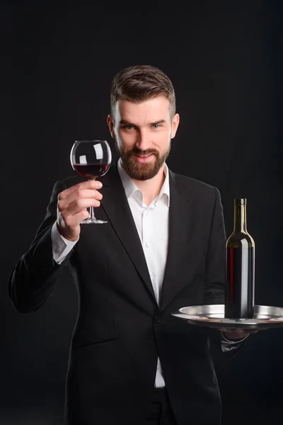 Sommelier raised his glass. Smiling bearded man holding a tray with a bottle of red wine. Vertical studio shot with black background.
