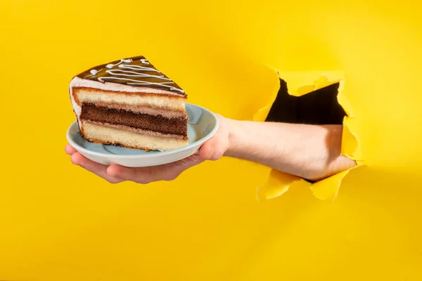 Mão segurando um grande pedaço de bolo de chocolate através de um buraco rasgado em papel amarelo — Fotografia de Stock