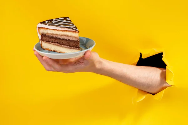 Mão oferecendo um pedaço de bolo através de um buraco rasgado no fundo de papel amarelo . — Fotografia de Stock