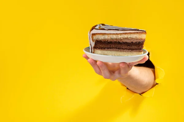Mão dando um pedaço de bolo de chocolate através de um buraco rasgado no fundo de papel amarelo . — Fotografia de Stock