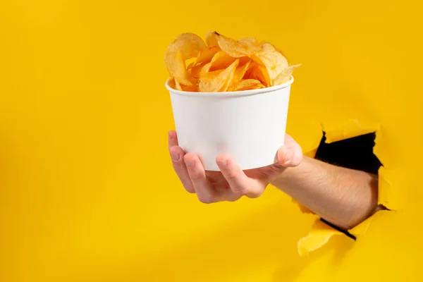 Mão dando uma xícara de batatas fritas através de um buraco rasgado em papel amarelo — Fotografia de Stock