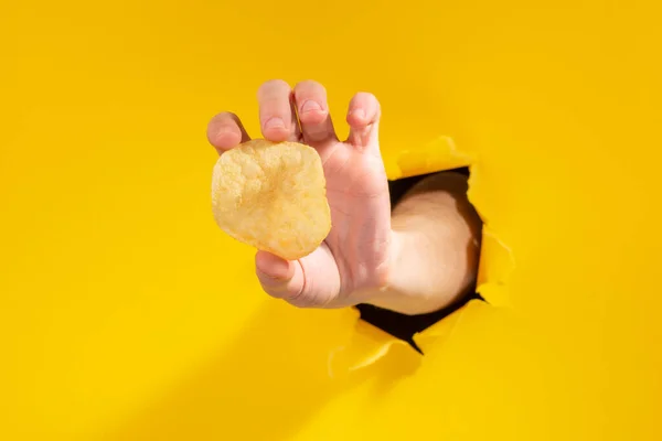 Mão segurando uma batata frita através de um buraco rasgado no fundo de papel amarelo . — Fotografia de Stock