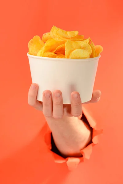 Mão segurando uma xícara inteira de batatas fritas através de um buraco rasgado no fundo vermelho — Fotografia de Stock