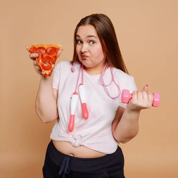 Niña gorda entrenando con mancuerna y sosteniendo pizza —  Fotos de Stock