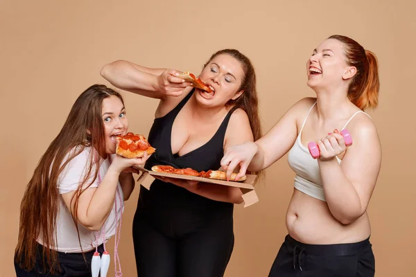 Três mulheres com excesso de peso comendo pizza em fundo bege — Fotografia de Stock