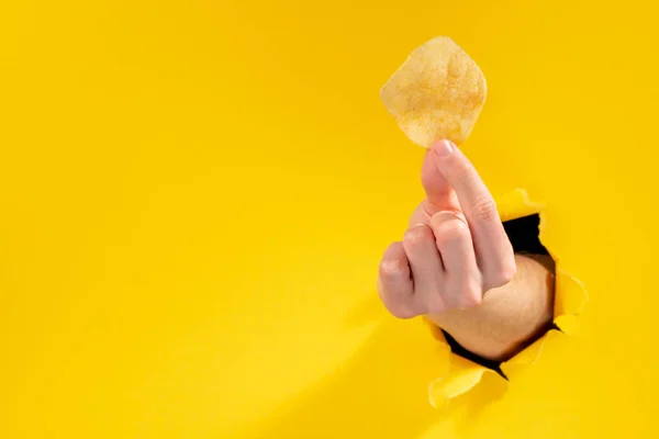 Mão segurando uma batata frita através rasgado fundo de papel amarelo — Fotografia de Stock
