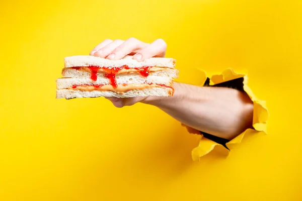 Mão segurando um sanduíche através de papel amarelo rasgado fundo — Fotografia de Stock