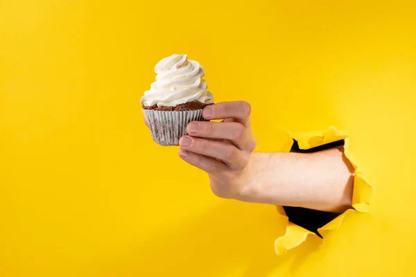 Main donnant un cupcake à travers un fond de papier jaune déchiré — Photo