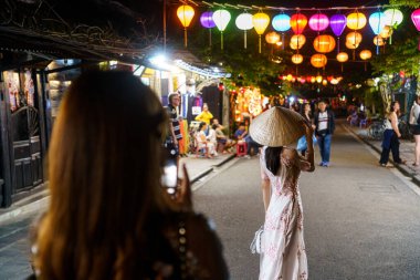 Bir gece sokakta beyaz Vietnam elbisesi giymiş bir kadının fotoğrafını çeken Asyalı bir kadın.