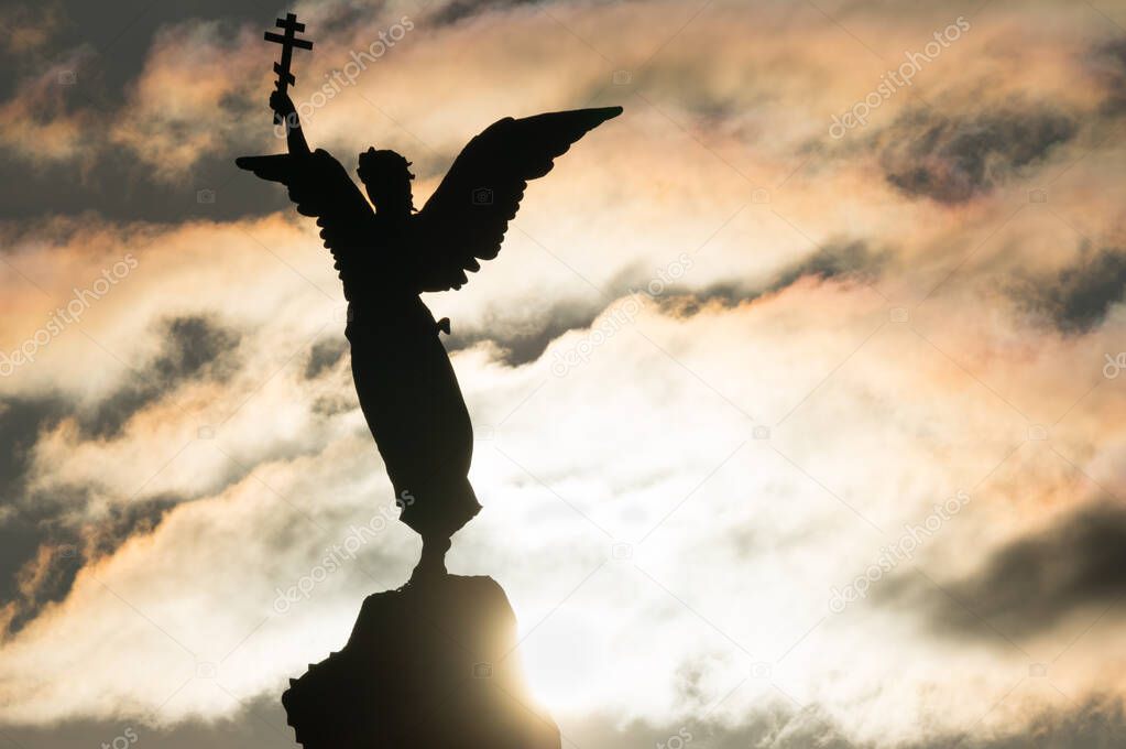 Silhouette of an angel with a cross in his hand on a background of dramatic sky and sun. Fragment of the Monument Mermaid , installed in 1902 in the Kadriorg park in Tallinn, Estonia