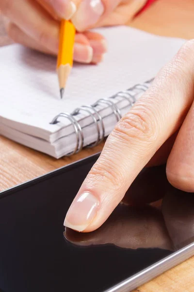 Hand of woman using mobile phone and writing notes in notepad — Stock Photo, Image