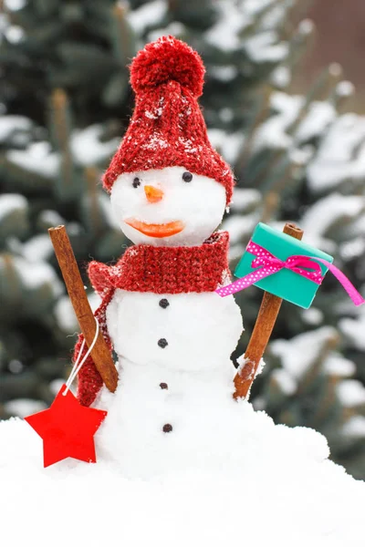 Inredda snögubbe med gåva till jul eller Valentine på bakgrund av barrträd omfattas snö — Stockfoto