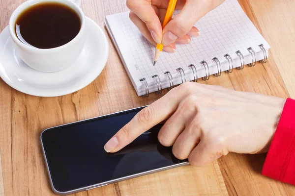 Hand of woman using mobile phone and writing notes in notepad, cup of coffee — Stock Photo, Image