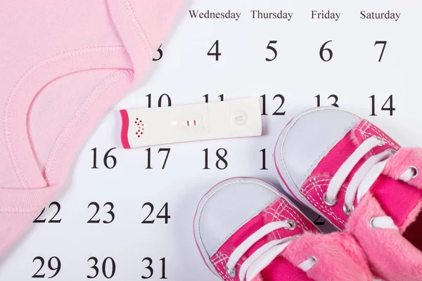 Test di gravidanza con risultato positivo e abbigliamento per neonato sul calendario, in attesa per il bambino — Foto Stock