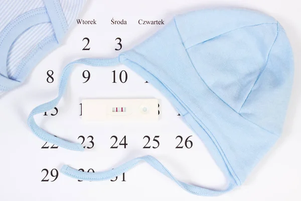 Pregnancy test with positive result and clothing for newborn on calendar, expecting for baby — Stock Photo, Image