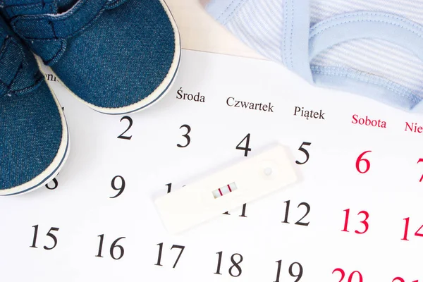 Pregnancy test with positive result and clothing for newborn on calendar, expecting for baby — Stock Photo, Image