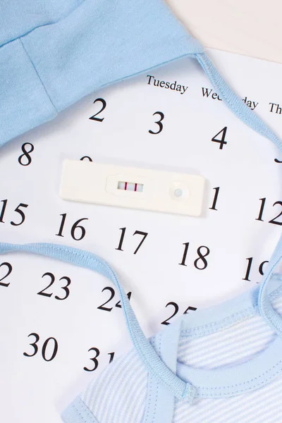 Pregnancy test with positive result and clothing for newborn on calendar, expecting for baby — Stock Photo, Image