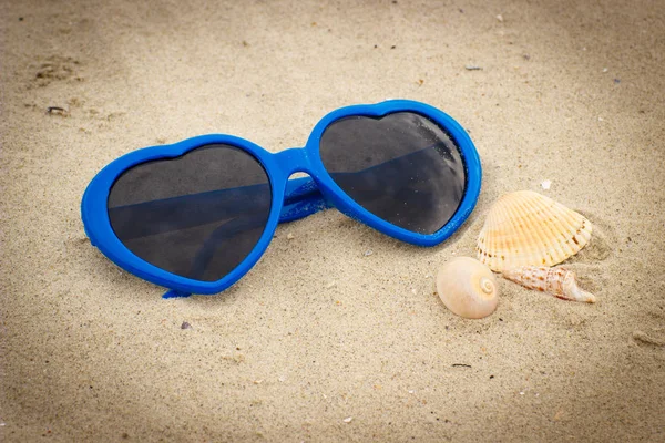 Occhiali da sole blu a forma di cuore con conchiglie sulla sabbia — Foto Stock