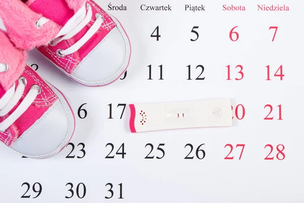Pregnancy test with positive result and baby shoes on calendar with polish inscription, expecting for baby concept — Stock Photo, Image
