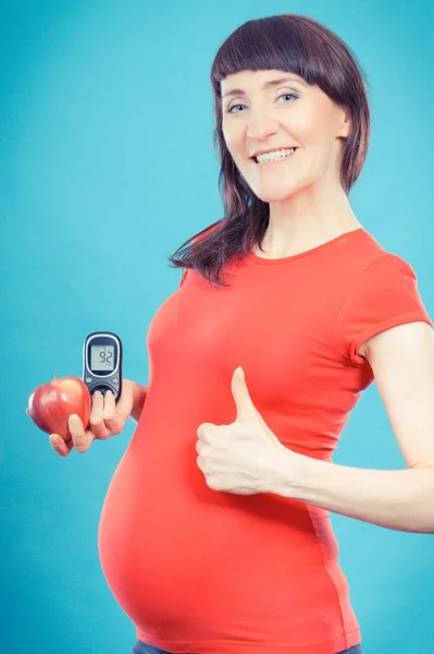 Счастливая беременная показывает большие пальцы, держа яблоко и глюкометр с хорошим результатом уровня сахара — стоковое фото