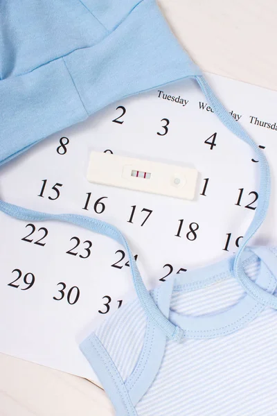 Test di gravidanza con risultato positivo e abbigliamento per neonato sul calendario, in attesa per il bambino — Foto Stock
