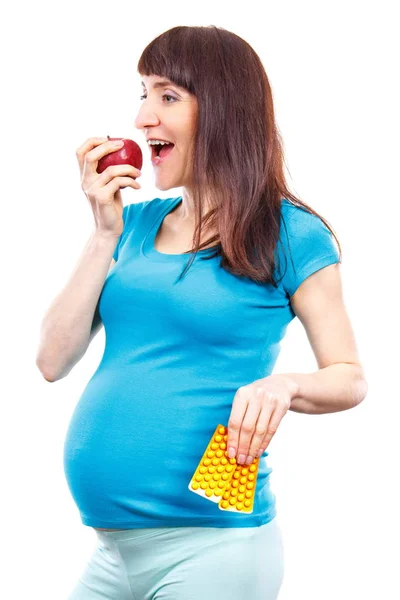 Щаслива вагітна жінка їсть свіже яблуко і тримає медичні таблетки або добавки, вибір між здоровою їжею і таблетками — стокове фото