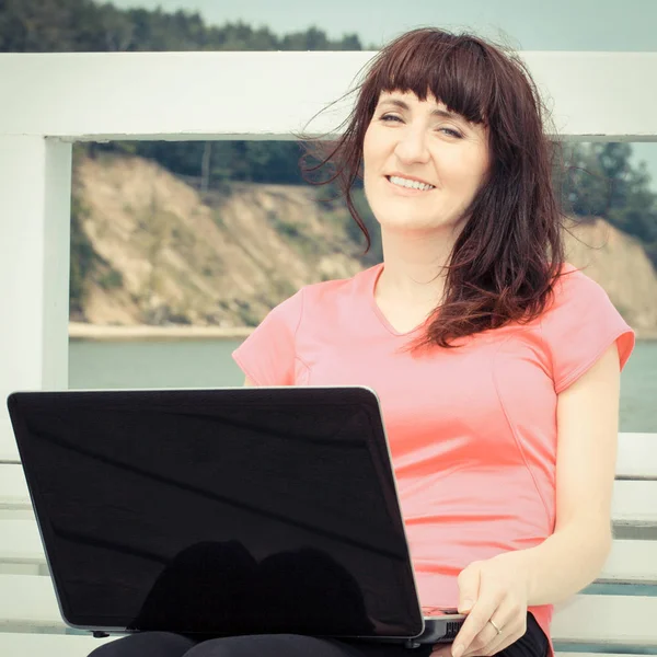 Винтажное фото, женщина сидит на скамейке у моря со своим ноутбуком, используя компьютерную концепцию — стоковое фото