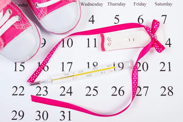 Test di gravidanza con esito positivo, termometro e scarpe da bambino sul calendario — Foto Stock