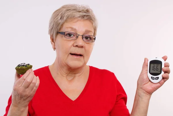 Verbaasd senior vrouw met glucose meter en verse cupcake, concept voor het meten en controleren van suikerniveau — Stockfoto