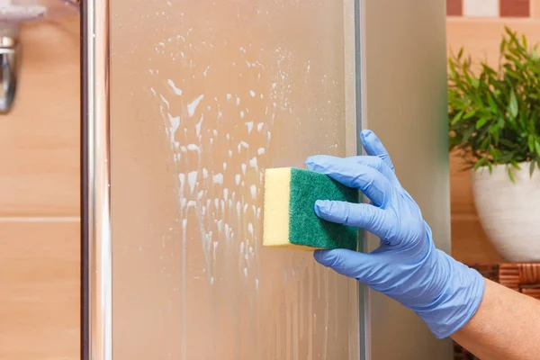 Hand of senior woman cleaning glass shower using detergent and sponge, household duties concept