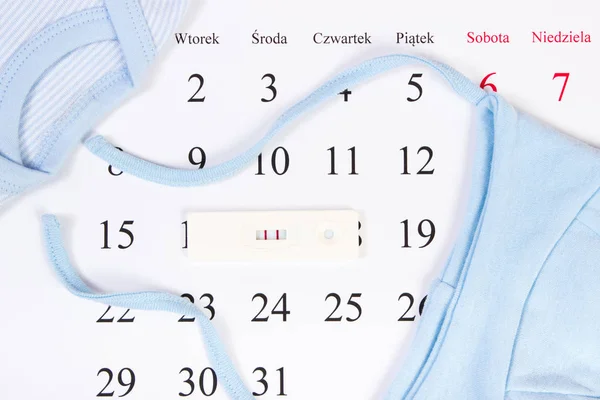 Test di gravidanza con risultato positivo e abbigliamento per il neonato sdraiato sul calendario, in attesa per il bambino — Foto Stock