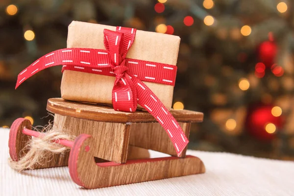 Τυλιγμένο δώρο με κορδέλα σε ξύλινο έλκηθρο και το χριστουγεννιάτικο δέντρο με φώτα στο παρασκήνιο — Φωτογραφία Αρχείου