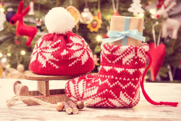 Wrapped gift for Christmas with woolen sock and cap on background of christmas tree with decoration — Stock Photo, Image