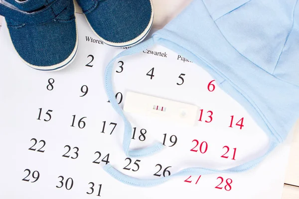 Test di gravidanza con risultato positivo e abbigliamento per neonato sul calendario — Foto Stock