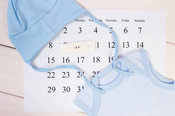 Test di gravidanza con risultato positivo e abbigliamento per il neonato sul calendario, in attesa del concetto di bambino — Foto Stock