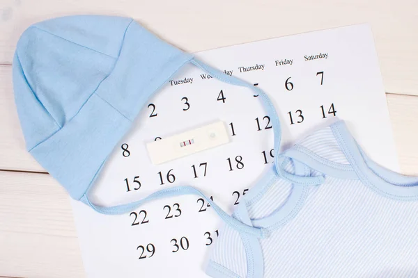 Test di gravidanza con risultato positivo e abbigliamento per il neonato sul calendario, in attesa del concetto di bambino — Foto Stock