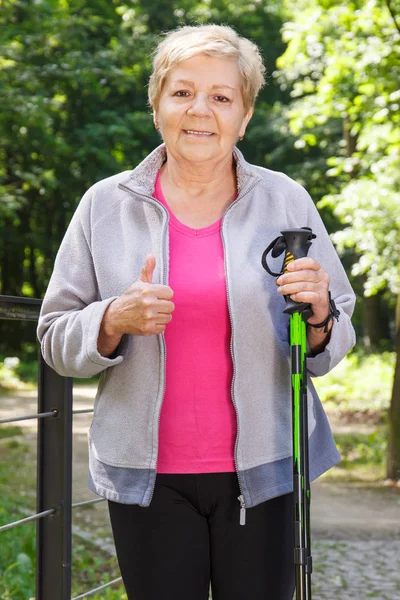 Starší vedoucí žena drží hole pro nordic walking a ukazuje palec nahoru — Stock fotografie