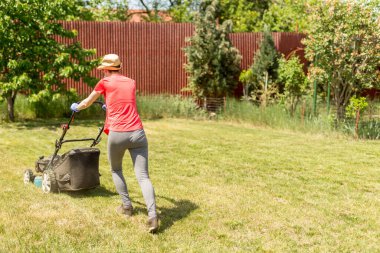 Bahçesinde çim biçme makinesiyle çim biçen bir kadın güneşli bir günde Coronavirus salgını yüzünden cerrahi maske takıyor..