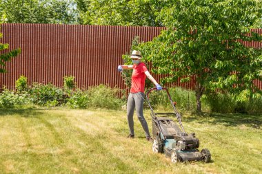 Bahçesinde çim biçme makinesiyle çim biçen bir kadın güneşli bir günde Coronavirus salgını yüzünden cerrahi maske takıyor..